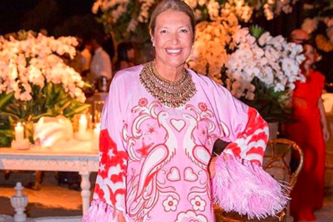Donata Meirelles, com um vestido rosa e estampado, canelos loiros presos e um colar. A diretora de estilo da Revista Vogue está em pé em um ambinete fechado com flores no fundo