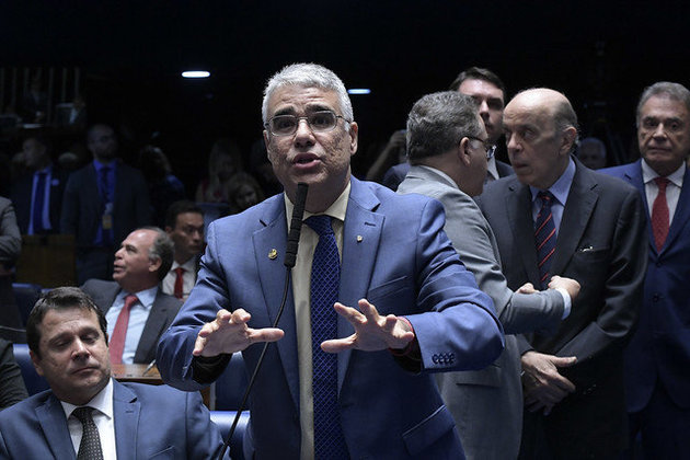 Senador Eduardo Girão, homem branco, 46 anos, de cabelos grisalhos, terno e gravata azul esccuro e camisa marrom clara. O senador está em pé, falando no microfone