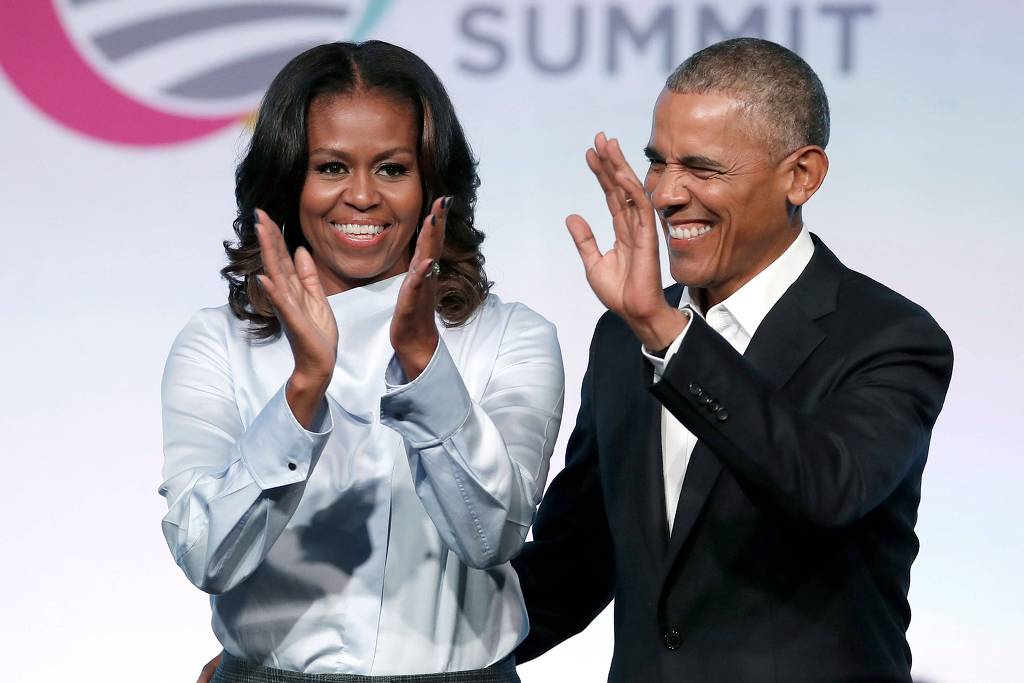 Michelle Obama- mulher negra, de cabelo liso, usando camiseta branca- e Barack Obama - homem negro, de pouco cabelo, usando terno- em pé acenando e sorrindo