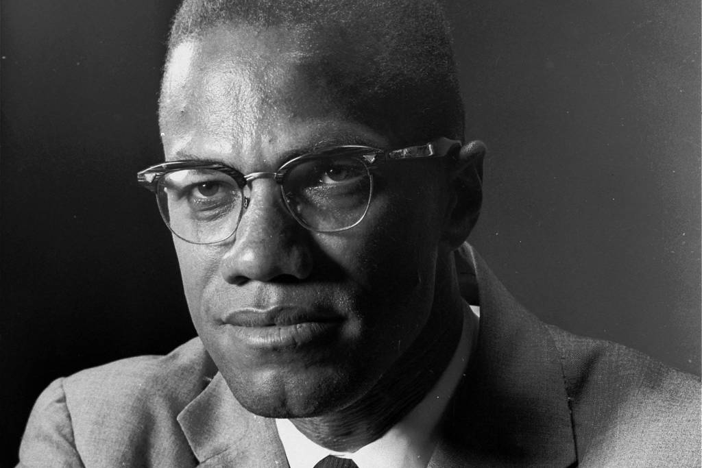 Malcolm X- homem negro, de pouco cabelo, usando óculos de grau, vestindo termo e gravata- sentado olhando para frente