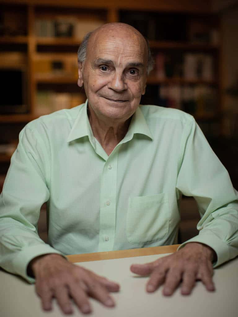 Foto de Drauzio Varella - homem branco idoso. calvo, usando camiseta de manga longa verde claro- sentado com as duas mãos em cima da mesa