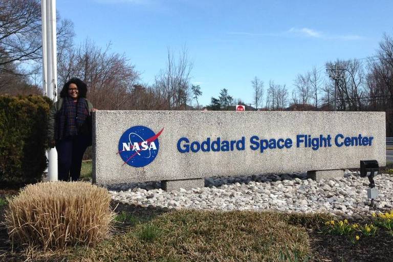 Carolyne Oliveira- mulher negra, de cabelo cacheado, usando roupas de frio- ao lado da placa do instituto da NASA