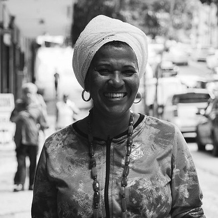 Foto em preto e branco de Makota Célia Gonçalves Souza- mulher negra, usando turbante- em pé sorrindo