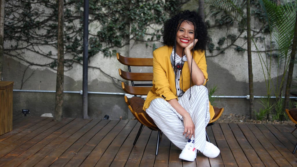 Samantha Almeida- mulher negra, de cabelo cacheado, usando macacão branco e blazer amarelo- sentada sorrindo