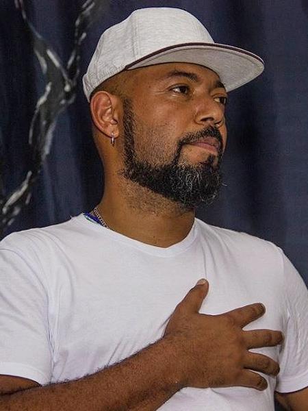 Douglas Belchior- homem negro, vestindo camiseta e boné branco- em pé com a mão no peito