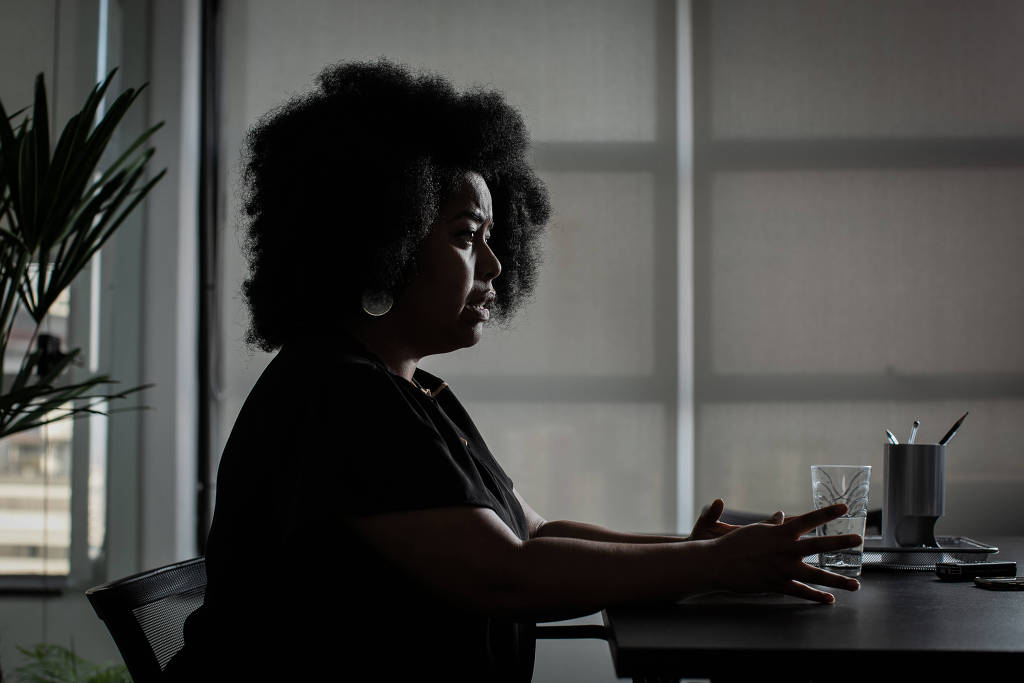 A advogada Silvia Souza- mulher negra, de cablo black power, vestindo camiseta preta- sentada em uma cadeira gesticulando