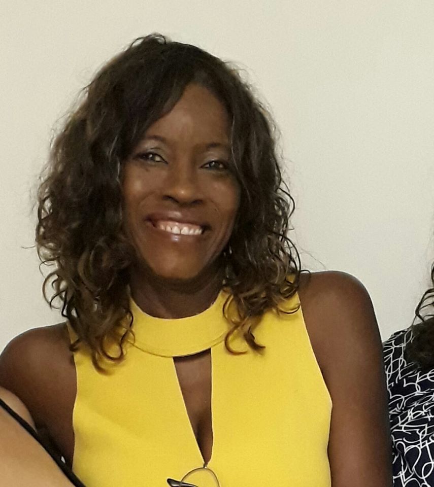 Eliane Leite - mulher negra de cabelo cacheada, vestindo camiseta amarela - em pé sorrindo.