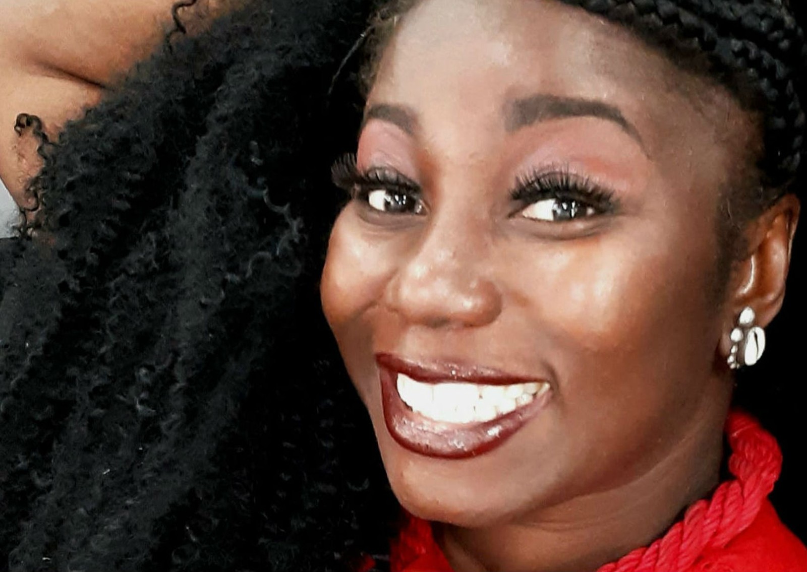Carla Akotirane - mulher negra d cabelo cacheado, usando camiseta e batom vermelho- sentada sorrindo