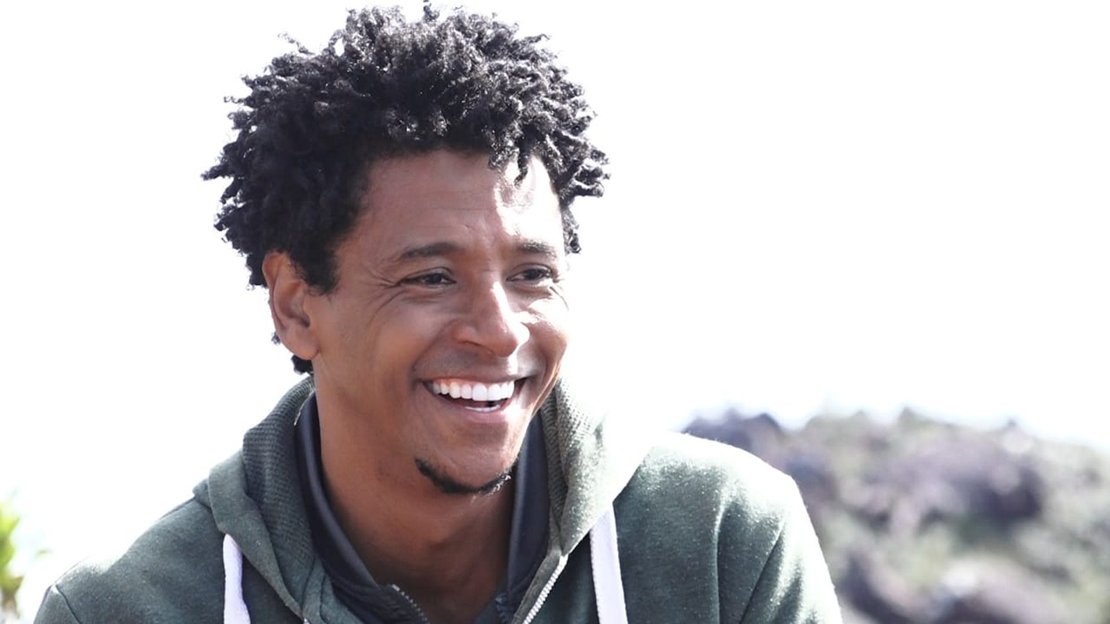 Aldri Nascimento - homem negro de cabelo crespo, usando moletom cinza- sentado ao ar livre sorrindo