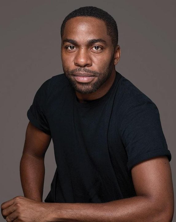 Lázaro Ramos - homem negro de pouca barba e cabelo, usando camiseta preta- sentado 
