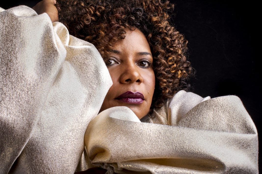 Margareth Menezes - mulher negra de cabelo cacheado, usando roupa beje- sentada com a mão esquerda apoiando o rosto