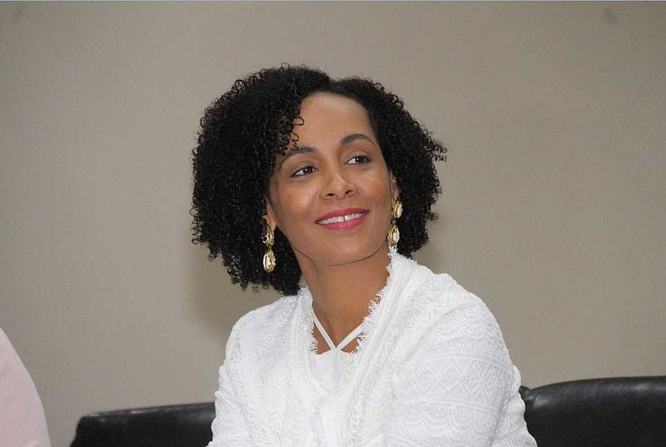 Lívia Vaz - mulher negra de cabelo cacheado, usando camisa branca e batom rosa- sentada sorrindo