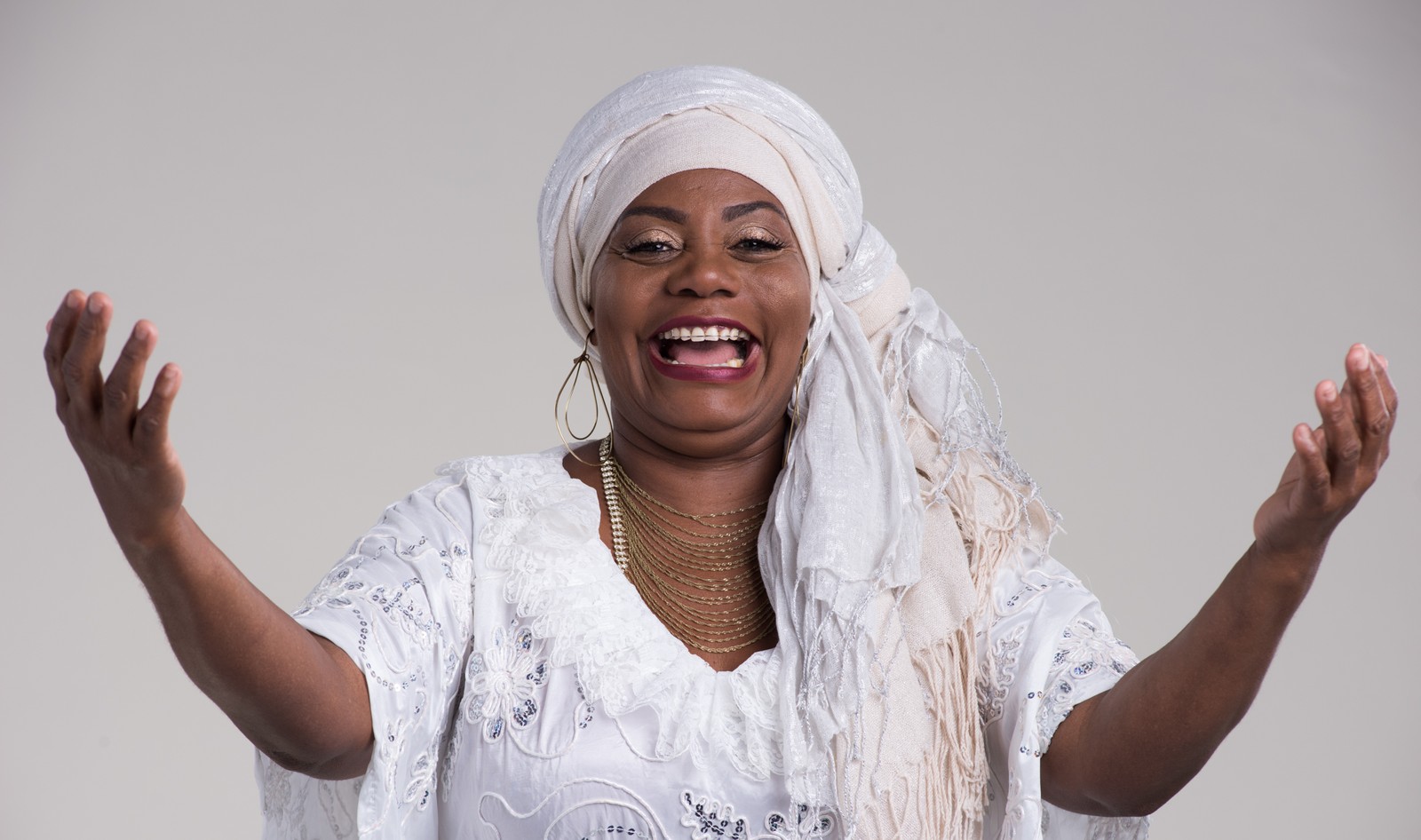 Dadá - mulher negra, usando turbante e roupa branca- em pé, sorrindo e com os braços estendido