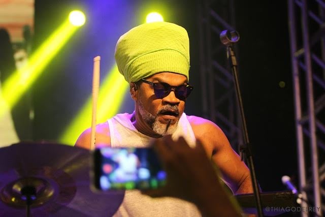 Carlinhos Brown - homem negro de barba grisalha, usando camiseta branca e turbante verde- tocando durante um show