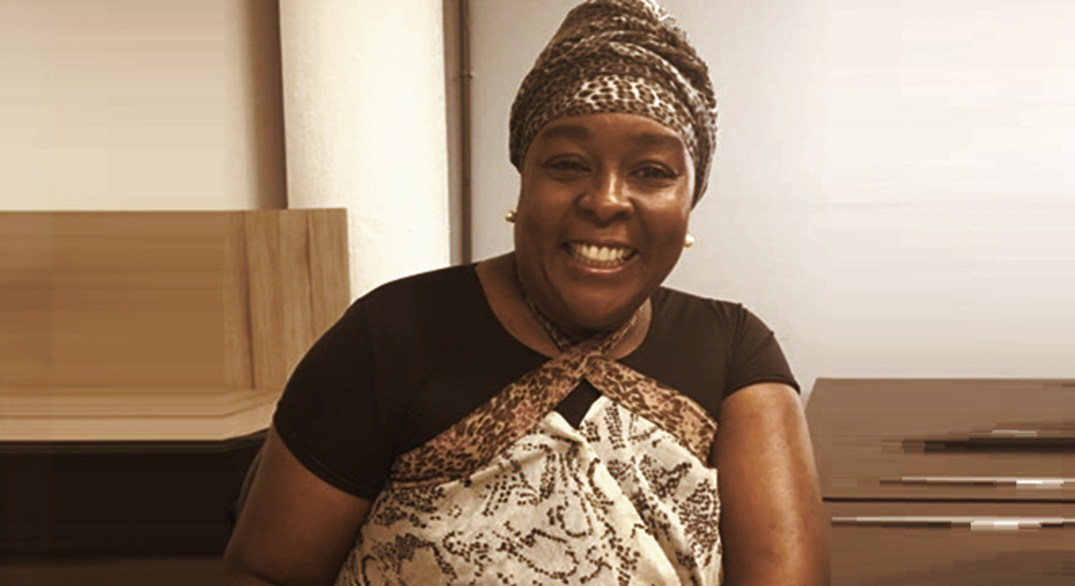 Regina Celia - mulher negra, usando turbante e blusa marrom - sentada sorrindo