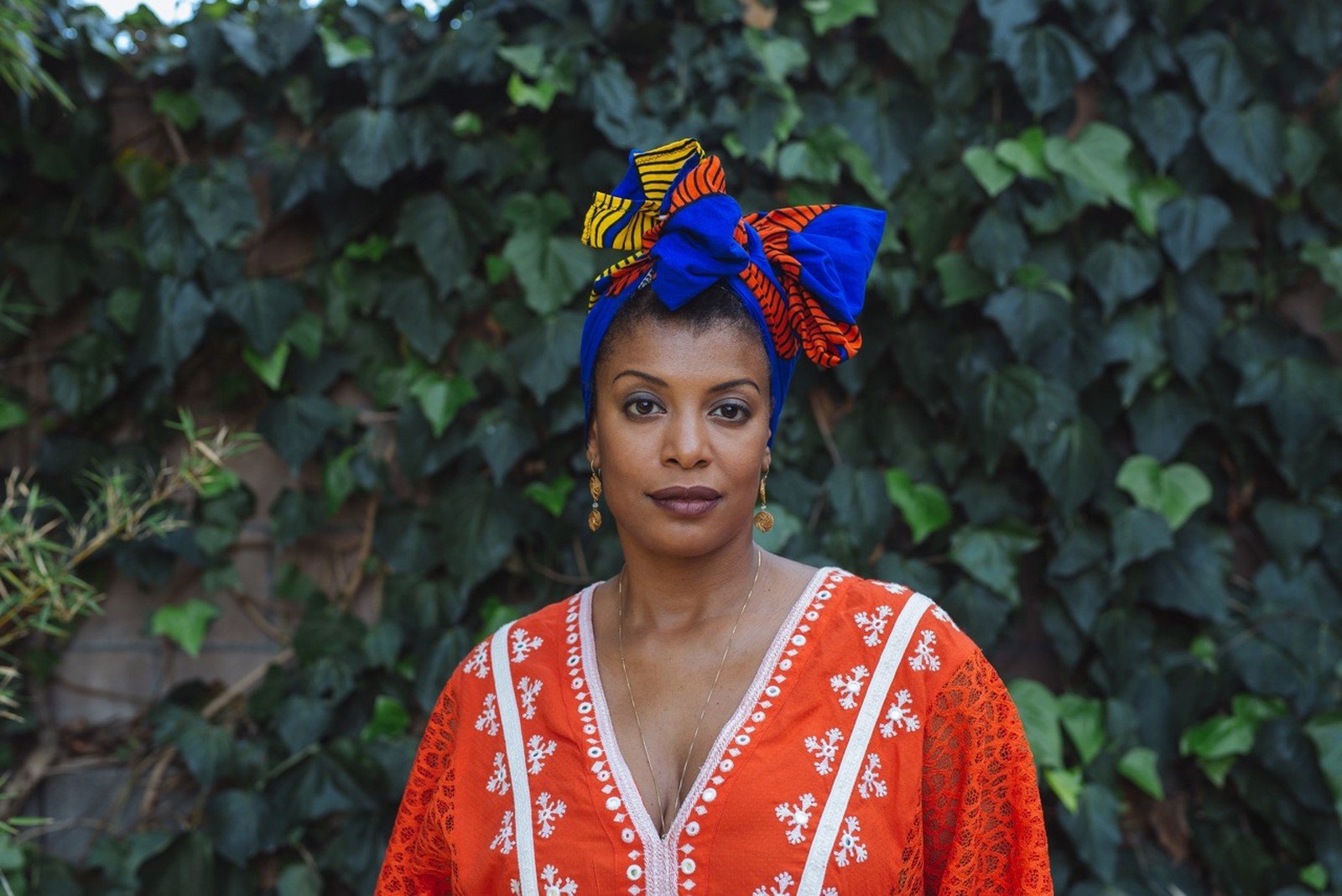 Thalma de Freitas - mulher negra, usando turbante azul marinho e camiseta laranja- em pé olhando para frente