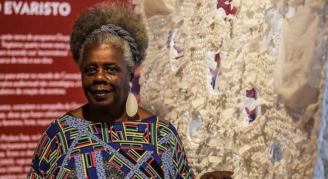 Conceição Evaristo- mulher idosa nera, de cabelo grisalho crespo, usando camiseta colorida e brincos grandes - em pé sorrindo