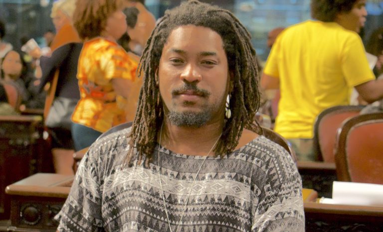 Jonathan Raymundo,- homem negro, de barba e dreads, vestindo camiseta estampada cinza- olhando para frente