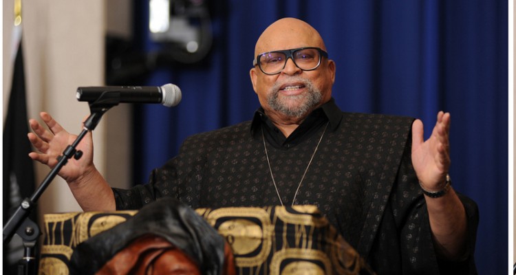Maulana Karenga- homem negro careca, usando camiseta preta e óculos de grau- falando e gesticulando com as mãos