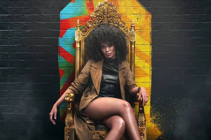Foto da atriz Pearl Thusi- mulher negra, de cabelo crespo, vestindo vestido preto e casaco marrom- sentada em uma cadeira