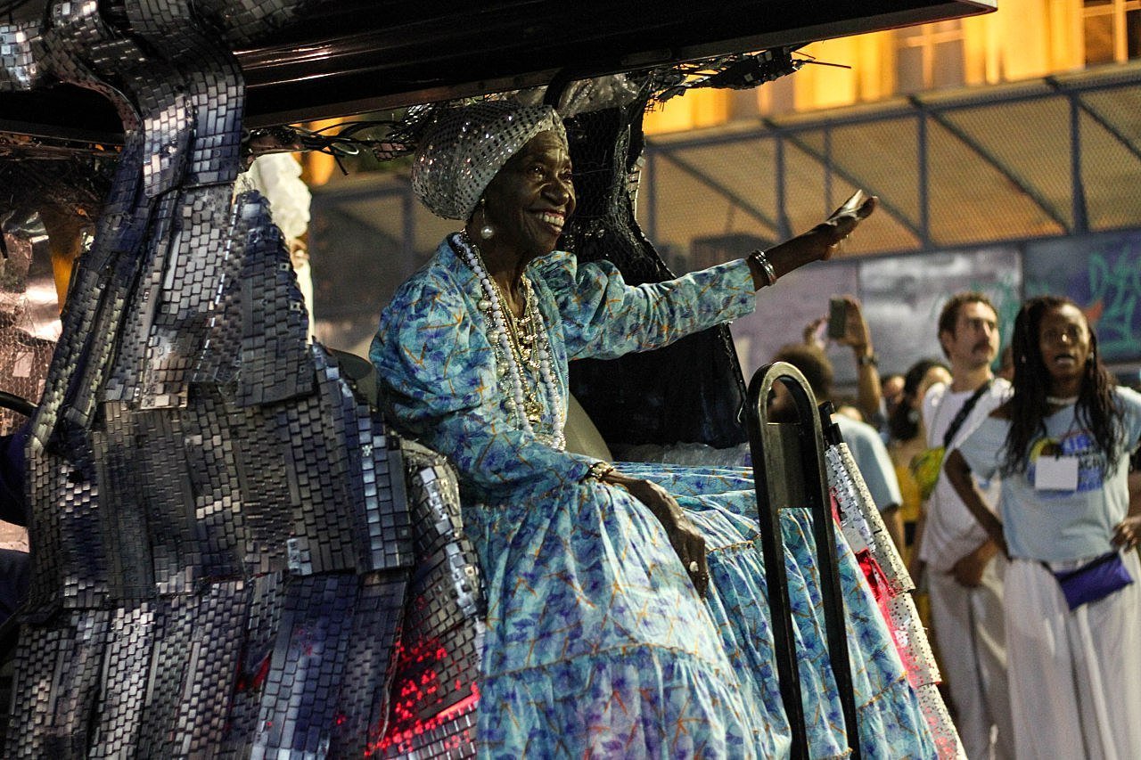 Lia de Itamaracá- mulher idosa negra, vestindo vestido azul, sentada em um carro alegórico