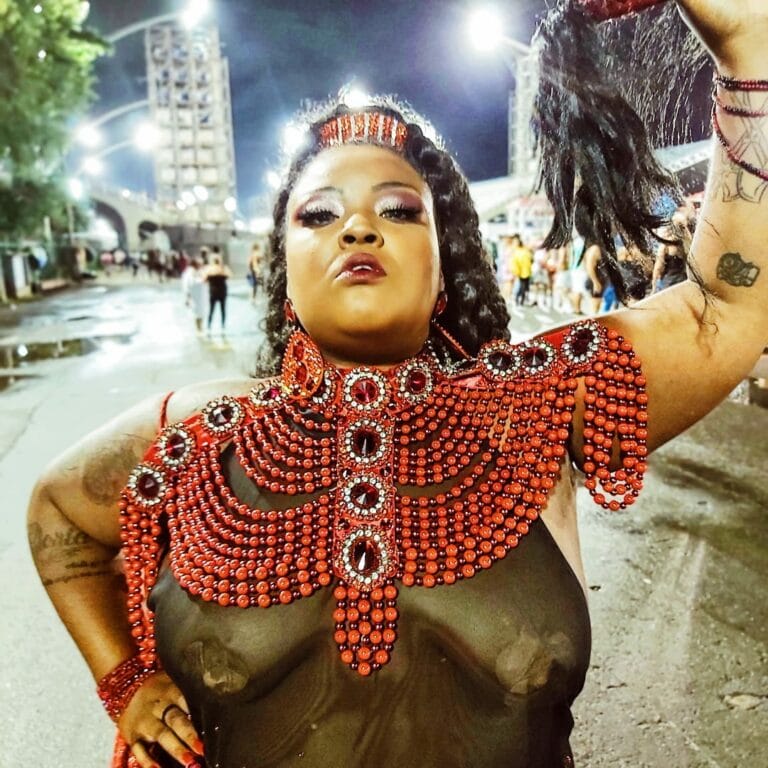 Joyce Carolina - mulher negra, de cabelo longo e ondulado, usando colar de pedras vermelhas- em pé