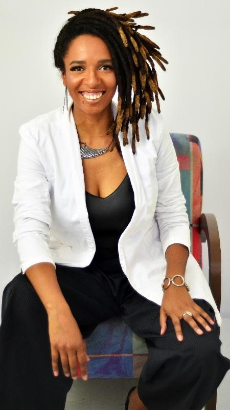 Camila de Moraes - mulher negra, com dreads, usando macacão preto e blazer branco- sentada em uma cadeira sorrindo