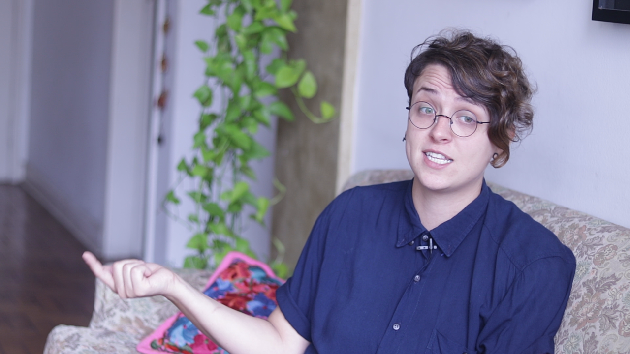 Debora Baldin- mulher branca de cabelo curto, usando óculos e camiseta azul marinho- sentada em um sofá