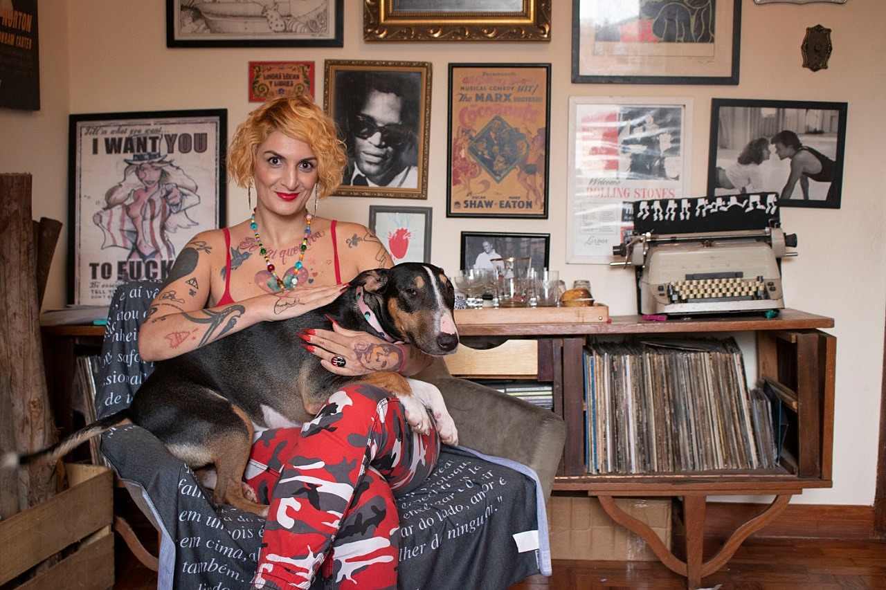 Clara Averbuck - mulher branca de cabelo loiro, usando mascação vermelho estampado- sentada em uma poltrona e segurando um cachorro no colo