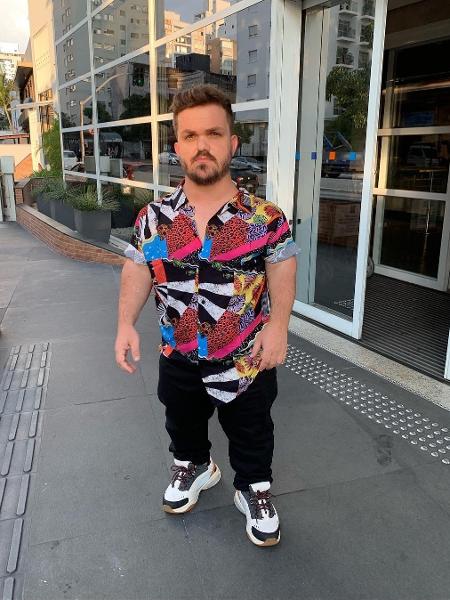 Mateus Baptistella- homem branco, de barba e cabelo longo, usando camiseta colorida e calça preta- em pé olhando para frente