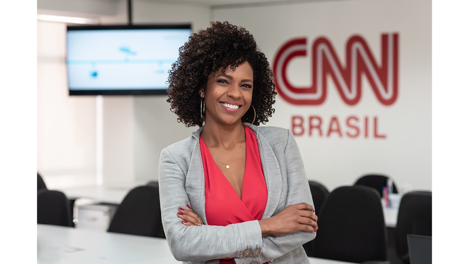 Luciana Barreto- mulher negra de cabelo cacheado, vestindo blusa rosa e blazer cinca- em pé, sorrindo e com os braços cruzados