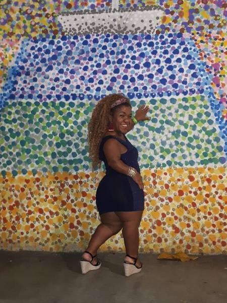 Vivi de Assis- mulher negra, de cabelo longo e cacheado, usando tamanco e vestido azul marinho- em pé sorrindo