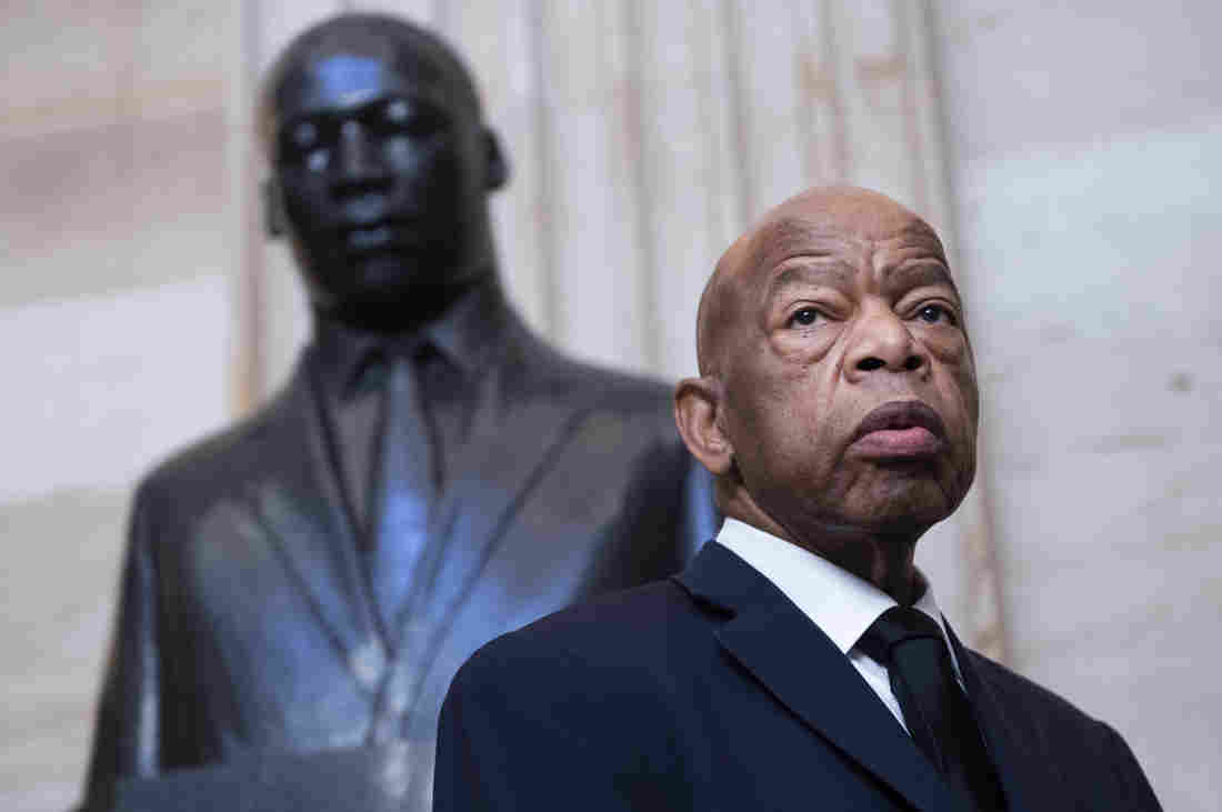 John Lewis- homem idoso nefro, careca, usando terno e gravata- em pé ao lado de uma estatua