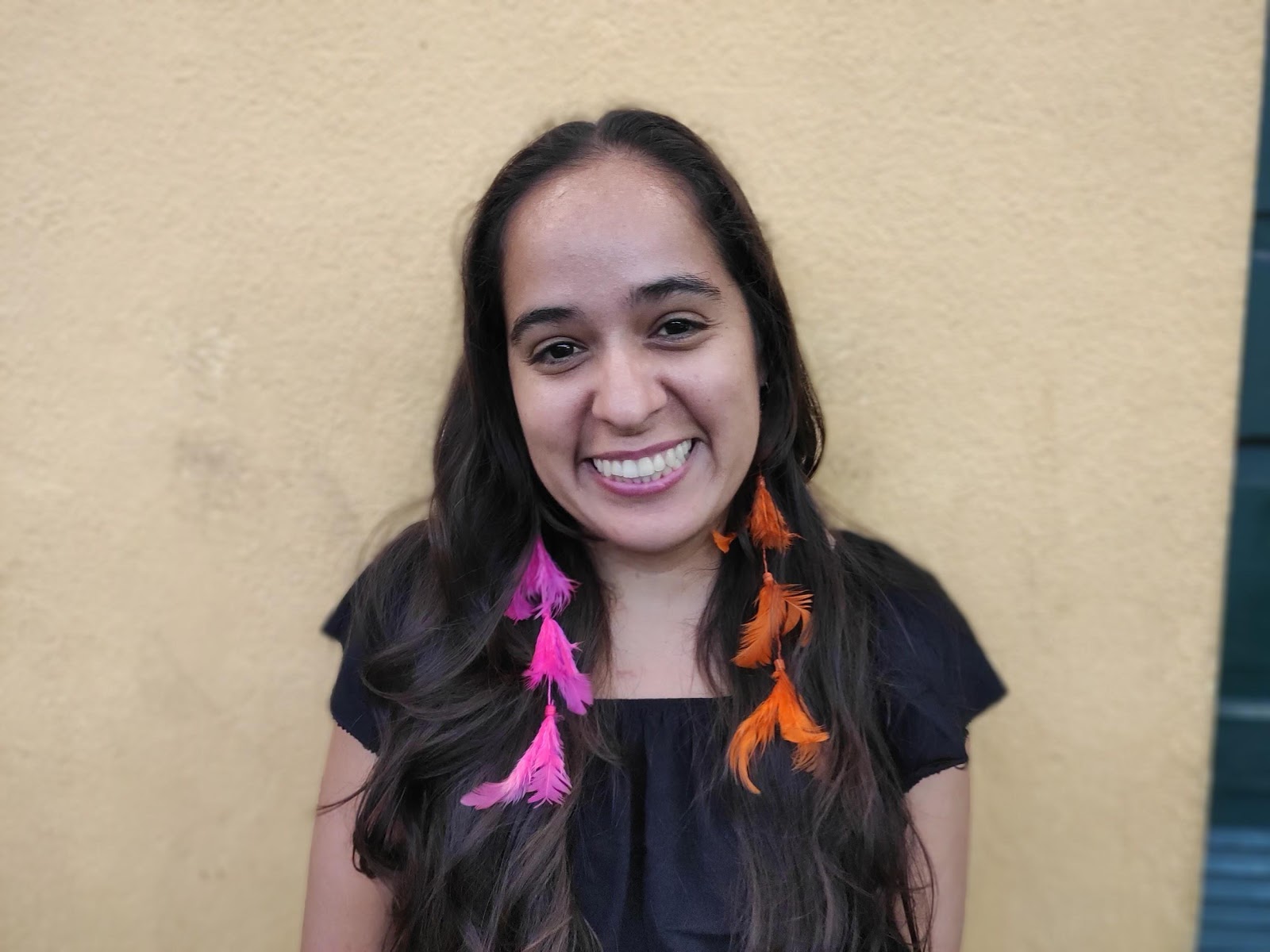 Geni Núñes,- mulher, de cabelo ondulado e longo, usando brinco de penas e camiseta preta- em pé sorrindo 