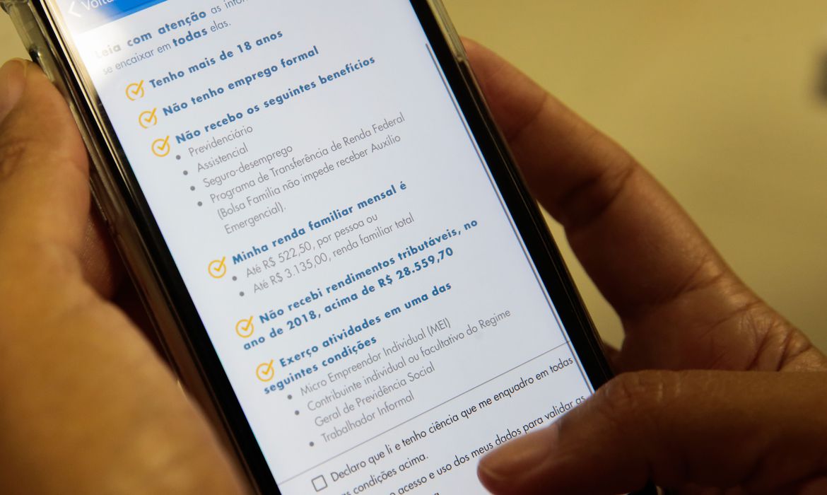 Foto de uma mão com segurando um celular, no celular mostra o app do auxilio emergencial